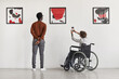 © Seventyfour - Back view portrait of young woman using wheelchair taking photo of artwork while visiting accessible museum, copy space