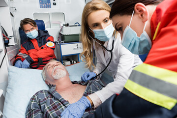 Selective focus of doctor with stethoscope looking at paramedic in medical mask and sick patient in ambulance car