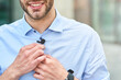 © Kostiantyn - Young handsome man, male blogger adjusting small lavalier microphone on his shirt and smiling while standing outdoors