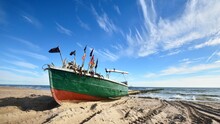 An Old Green Fishing Boat On The Sandy Shore Close-up, Motorboat Moored To A Pier In The Background. Clear Blue Sky. Baltic Sea, Latvia