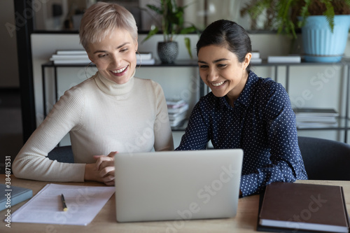 Smiling multiracial female colleagues sit at desk in office look at laptop screen watch funny video at workplace. Happy diverse multiethnic coworkers have fun using computer enjoy break in office.
