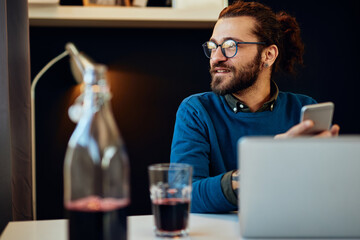 Wall Mural - Young smiling handsome bearded businessman sitting in his office and taking a break. He is typing message on mobile.