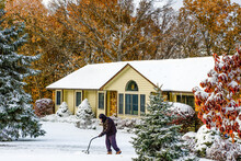 Midwestern House And Landscape Around It Covered With Early Snow; Trees Still Have Fall Foliage; Man In Warm Clothes Shoveling Driveway; Winter In Missouri