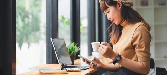 Beautiful asian businesswoman in the cafe, using smartphone and drinking coffee smiling.Focus on coffee cup,Soft focus.