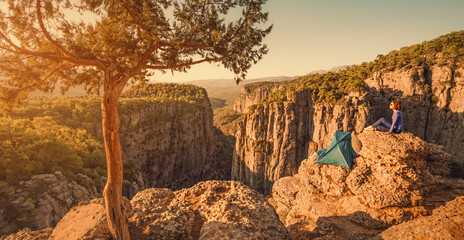 Wall Mural - Woman hiker has set up a tent on top of a cliff overlooking a deep gorge and beautiful mountains during bright sunrise. Hiking and rock climbing in a natural national Park