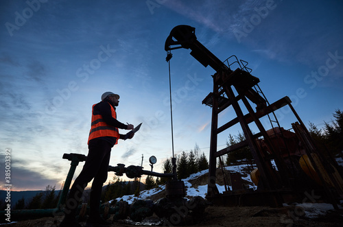 Low angle side view snapshot of petroleum engineer wearing orange vest and a helmet, looking at the oil rig and making some notes against beautiful evening sky, sunset