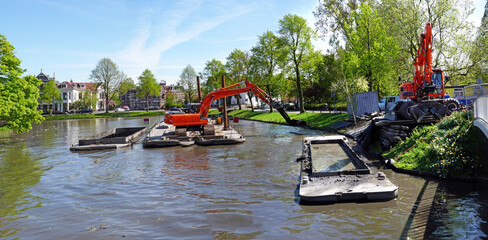 Dredging a city canal by crane from a pontoon; dredging spoil is transported in barges to a transfer site. 