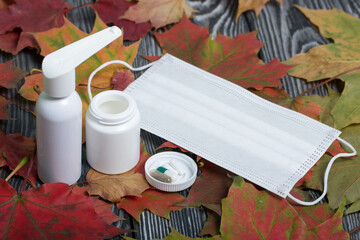 Wall Mural - Medical mask, container with pills and other drugs. The lid of the container and several tablets are next to each other. Among the autumn maple leaves on the black pine boards.