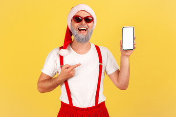 Wall Mural - happy smiling man with gray beard in sunglasses and santa claus hat pointing finger at smartphone wi