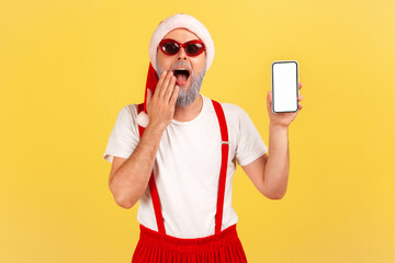 Wall Mural - shocked surprised man in stylish sunglasses and santa claus red hat holding smartphone with empty sc
