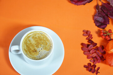 White cup of coffee with cappuccino cream on an orange background with autumn leaves, apples and red berries. Fall season, leisure time and coffee break concept. top view.