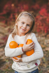 Wall Mural - Portrait of a beautiful little blonde girl in white christmas sweater holding a lot of oranges fruits and smiling in the park on autumn background