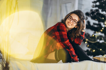 Pretty woman in shirt and socks lying in bed with christmas gift box, new year tree behind.