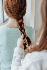 Two girls braid their hair at the window. Woman makes a braid to her friend. Hair weaving hairstyles. Girlfriend braids her hands with ringlets. Hair care