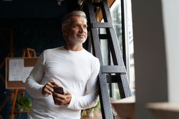 Business lifestyle. Casual Grey-haired Mature professional handsome businessman standing near window in his office while thinking about his goals