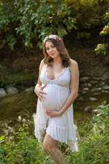 Close up portrait of a beautiful young expecting mother hands on her pregnancy belly wearing white pure dress flowers in her hair 