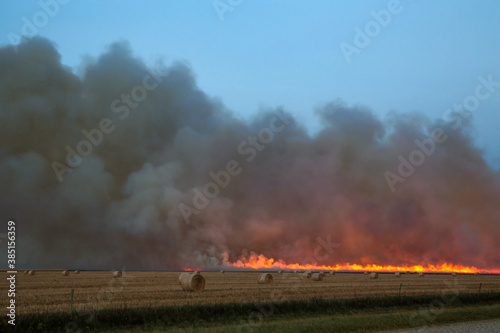 A line of fire with billowing smoke behind a field dotted with straw bales in an autumn evening landscape