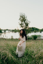 Brunette Filipino Woman Standing In A Grassy Field Near A Pond At Sunset Wearing A Tan Striped Belted Dress