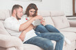 © ruslanshug - beautiful young couple family male and female using tablet pad device sitting on sofa indoor home room