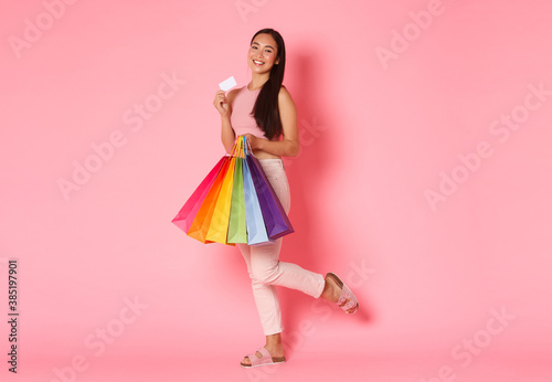Fototapeta Full length portrait of upbeat and excited, smiling trendy asian girl showing her credit card and looking pleased, carry shopping bags, bought new clothes for vacation, pink background