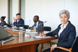 © AnnaStills - Portrait of mature businesswoman looking at camera while sitting at conference with her colleagues in the background