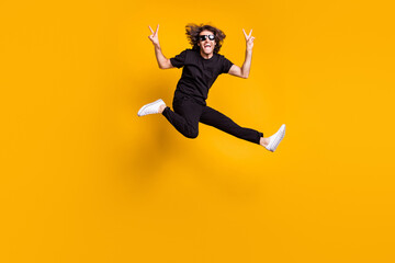 Full length body size portrait of young man jumping happily wearing black clothes demonstrating v-sign gesture with both hands isolated on bright yellow color background