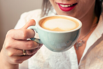  Woman is smiling and holding a cup of coffee. Hand with mug of coffee in cafe. Close up. Heart made from milk.