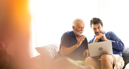 Wall Mural - Father and son family time together at home concept. Smiling old father and happy son sitting on sofa using digital laptop computer in living room at home