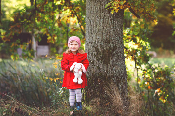 Beautiful toddler girl in red coat making a walk through autumn forest. Happy healthy baby holding soft toy rabbit. Sunny fall day with child. Active leisure and activity with kids in nature.