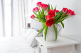 Fototapeta Tulipany - Bedroom in soft light colors..White vase with red tulips in light cozy bedroom interior. White wall, bed with white linen,