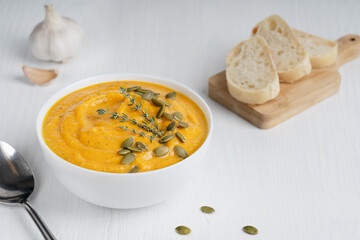 Wall Mural - Homemade vegan organic pumpkin soup made of pureed vegetables decorated with seeds and fresh thyme served in white bowl with bread slices, spoon and garlic on wooden background. Horizontal orientation