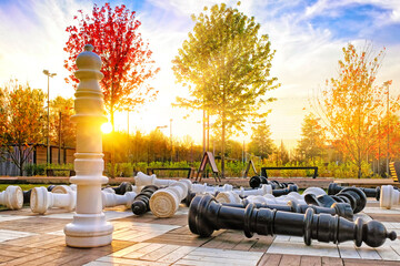 Wall Mural - sport playground in park with giant chess figures on checker board to play outdoor chess against golden autumn sunset background. Fallen chess figures