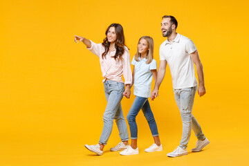 Wall Mural - Full length portrait of smiling funny young parents mom dad with child kid daughter teen girl in t-shirts holding hands pointing index finger aside isolated on yellow background. Family day concept.
