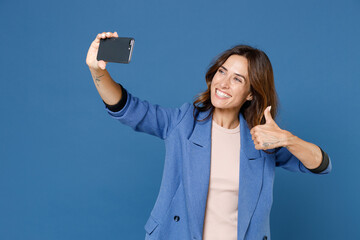 Wall Mural - Smiling cheerful attractive beautiful young brunette woman 20s wearing basic jacket doing selfie shot on mobile phone showing thumb up isolated on bright blue colour wall background, studio portrait.