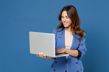 Smiling cheerful funny beautiful attractive young brunette woman 20s wearing basic casual jacket standing working on laptop pc computer isolated on bright blue colour background studio portrait.