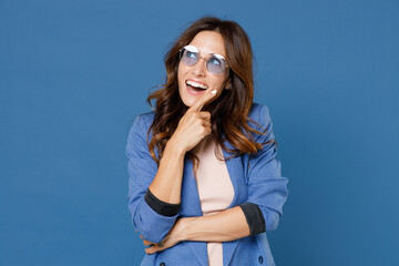 Wall Mural - Cheerful funny excited pensive young brunette woman 20s wearing basic jacket eyeglasses standing put hand prop up on chin looking aside up isolated on bright blue colour background studio portrait.