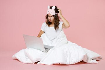 Wall Mural - Puzzled young woman in pajamas home wear sleep mask sitting working on laptop pc computer put hand on head while resting at home isolated on pastel pink background studio. Relax good mood concept.