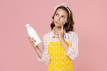 Pensive Young Woman Housewife 20s In Yellow Apron Checkered Shirt Hold Bottle Of Milk Put Hand Prop Up On Chin Doing Housework Isolated On Pastel Pink Background Studio Portrait. Housekeeping Concept.