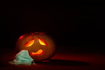 .Halloween pumpkin with sad scared face and coronavirus protective surgical mask next to it. New normal halloween on dark background