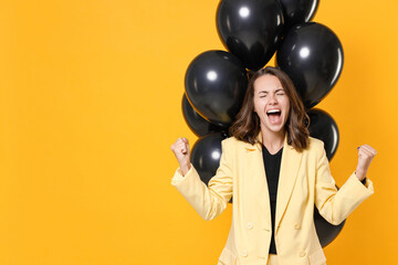 Screaming young woman in suit jacket celebrating birthday holiday party with bunch of air ballons clenching fists doing winner gesture isolated on yellow background studio portrait. Black friday sale.