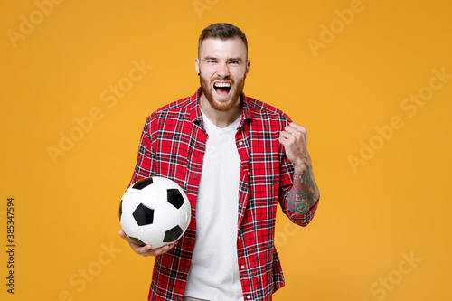 Screaming young man football fan in basic shirt cheer up support favorite team with soccer ball clenching fist doing winner gesture isolated on yellow background studio. People sport leisure concept.