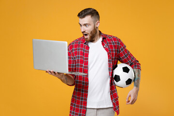 Shocked young man football fan in basic red checkered shirt cheer up support favorite team with soccer ball using laptop pc computer isolated on yellow background studio. People sport leisure concept.