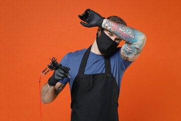 Exhausted professional tattooer master artist tattooed man in t-shirt apron face mask hold machine black ink jar equipment for making tattoo art on body put hand on head isolated on brown background.