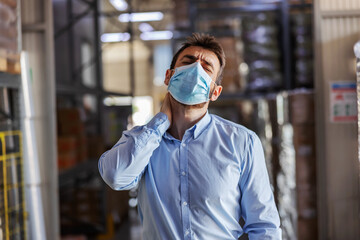 Young attractive businessman with face mask on having neck pain. Warehouse interior. Corona outbreak concept.