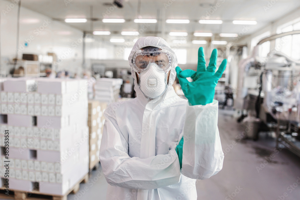 Worker in sterile uniform with rubber gloves holding sprayer with disinfectant and spraying around warehouse. Corona outbreak concept. - obrazy, fototapety, plakaty 
