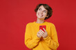 © ViDi Studio - Smiling pensive young brunette woman 20s wearing casual yellow sweater standing using mobile cell phone typing sms message looking aside up isolated on bright red colour background studio portrait.