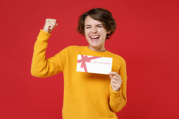 Wall Mural - Happy joyful young brunette woman 20s wearing basic yellow sweater standing hold in hands gift certificate doing winner gesture looking camera isolated on bright red colour background studio portrait.