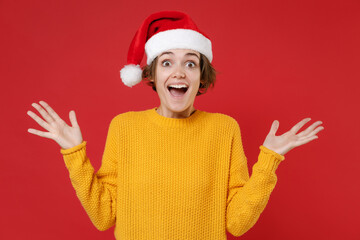 Canvas Print - Amazed excited young Santa woman in basic yellow sweater Christmas hat keeping mouth open spreading hands isolated on red background studio portrait. Happy New Year celebration merry holiday concept.