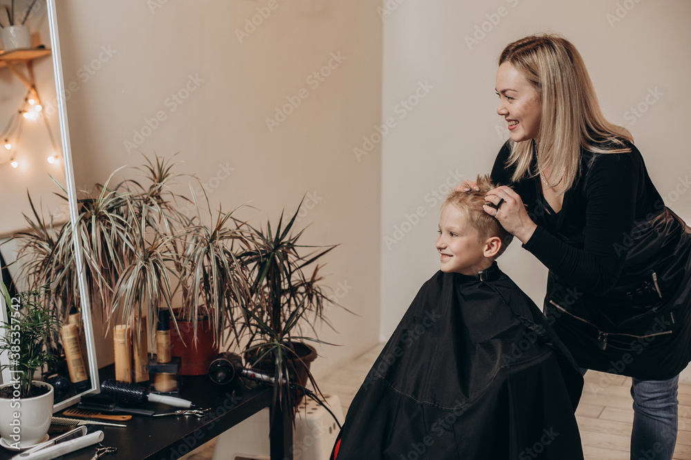 Eight-year-old blonde boy at the barbershop. A hairdresser cuts a haircut at a home beauty salon. Selective focus. - obrazy, fototapety, plakaty 