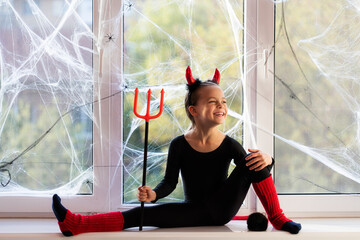 Wall Mural - Little girl gymnast in costume of little devil smiling and stretching on window sill